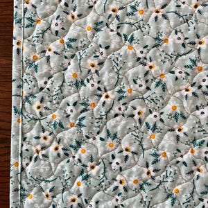 Pu&ograve; includere: Primo piano di un sottopiatto trapuntato con motivo floreale. Il tessuto &egrave; di colore verde-azzurro chiaro, ricoperto di fiori bianchi con centri gialli e foglie verdi. I bordi sono accuratamente cuciti con un bordo bianco.