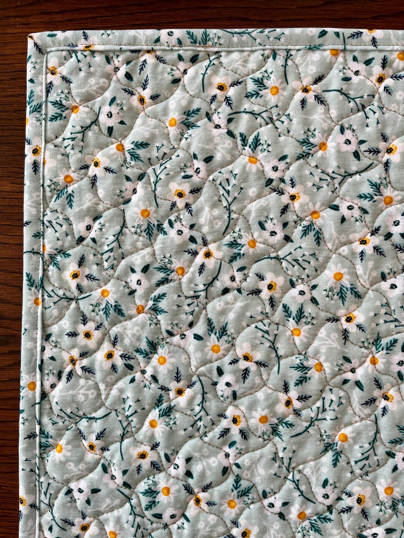 Pu&ograve; includere: Primo piano di un sottopiatto trapuntato con motivo floreale. Il tessuto &egrave; di colore verde-azzurro chiaro, ricoperto di fiori bianchi con centri gialli e foglie verdi. I bordi sono accuratamente cuciti con un bordo bianco.