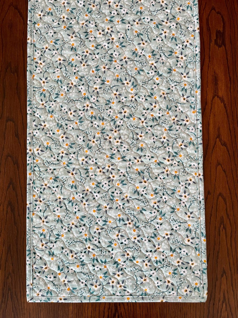 Pu&ograve; includere: Runner da tavolo trapuntato rettangolare con motivo floreale. Il tessuto &egrave; di colore verde-azzurro chiaro, ricoperto da piccoli fiori bianchi, centri gialli e foglie verdi. I bordi sono rifiniti con un bordo bianco.