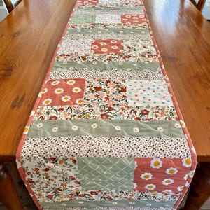 May include: A patchwork table runner with floral patterns in coral, green, and white. The runner is placed on a wooden table. The runner's design includes various floral patterns. The table runner is approximately 183 cm long, showcasing its decorative appeal.