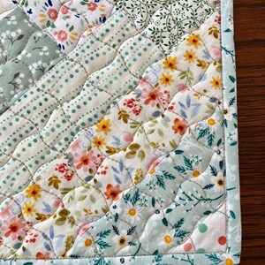 Pu&ograve; includere: Primo piano di un sottopiatto trapuntato con una variet&agrave; di motivi floreali in colori pastello. Il design include file diagonali di tessuto con fiori, pois e foglie. I bordi sono rifiniti con un bordo azzurro.