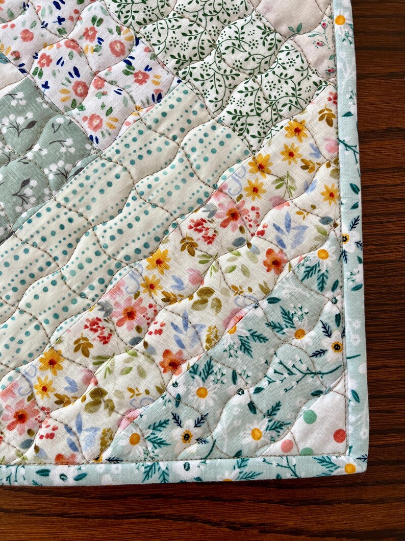 Pu&ograve; includere: Primo piano di un sottopiatto trapuntato con una variet&agrave; di motivi floreali in colori pastello. Il design include file diagonali di tessuto con fiori, pois e foglie. I bordi sono rifiniti con un bordo azzurro.