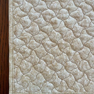 May include: Close-up of a quilted fabric featuring a paisley pattern in beige and cream hues. The fabric has a diamond-shaped quilted design. The edges are finished with a cream-coloured border. The fabric is likely used for home decor items.