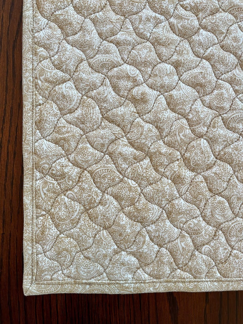 May include: Close-up of a quilted fabric featuring a paisley pattern in beige and cream hues. The fabric has a diamond-shaped quilted design. The edges are finished with a cream-coloured border. The fabric is likely used for home decor items.