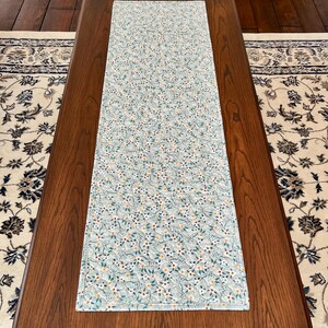 Pu&ograve; includere: Una tovaglia rettangolare con un motivo floreale azzurro. La tovaglia &egrave; posizionata su un tavolo di legno marrone scuro, con un tappeto fantasia visibile sullo sfondo. Il design floreale comprende fiori bianchi, foglie verdi e piccoli accenti arancioni.