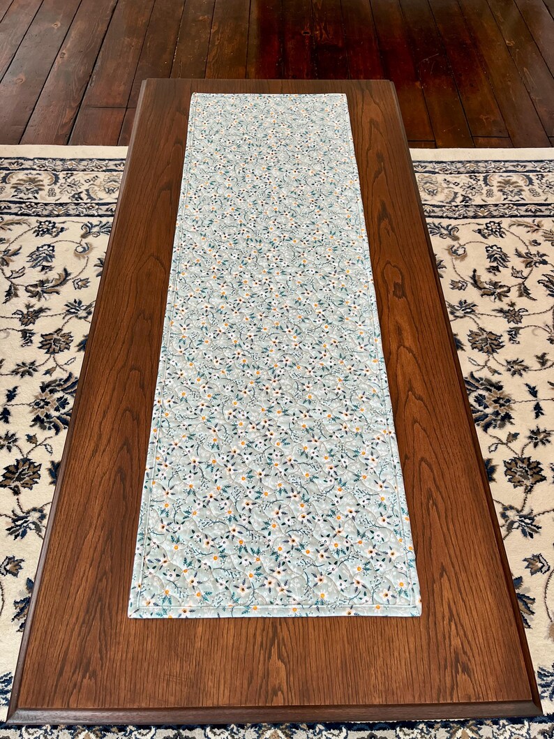 Pu&ograve; includere: Una tovaglia rettangolare con un motivo floreale azzurro. La tovaglia &egrave; posizionata su un tavolo di legno marrone scuro, con un tappeto fantasia visibile sullo sfondo. Il design floreale comprende fiori bianchi, foglie verdi e piccoli accenti arancioni.