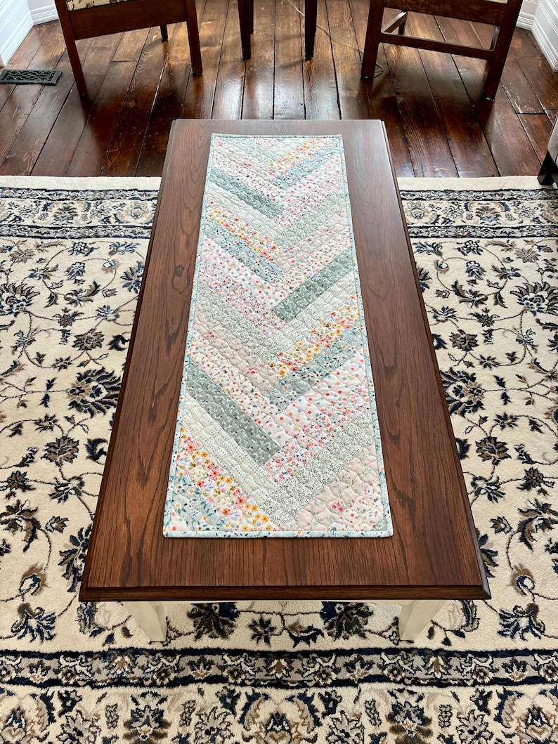 Pu&ograve; includere: Runner da tavolo trapuntato con motivo a spina di pesce. Il runner presenta un design patchwork con colori pastello, tra cui azzurro, rosa e stampe floreali. &Egrave; posizionato su un tavolino da caff&egrave; in legno marrone scuro.