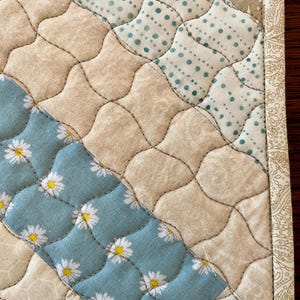 May include: Close-up of a quilted placemat with a patchwork design. The placemat features sections of beige, light blue with white daisy patterns, and light blue with white polka dots. The edges are trimmed with a patterned border.