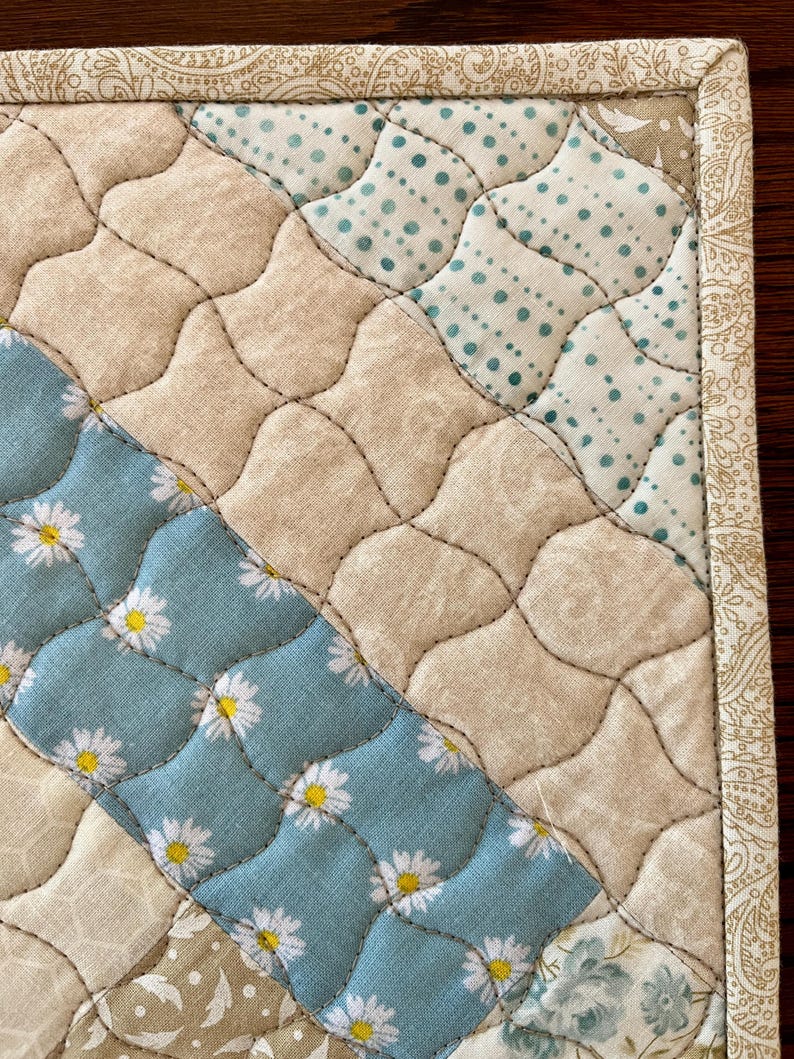 May include: Close-up of a quilted placemat with a patchwork design. The placemat features sections of beige, light blue with white daisy patterns, and light blue with white polka dots. The edges are trimmed with a patterned border.