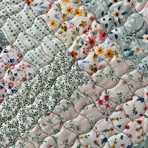 Pu&ograve; includere: Primo piano di un tessuto trapuntato con un design patchwork. La trapunta presenta vari motivi floreali nei toni del rosa, giallo, blu e verde su sfondo bianco. Le cuciture creano un motivo diagonale strutturato sul tessuto.