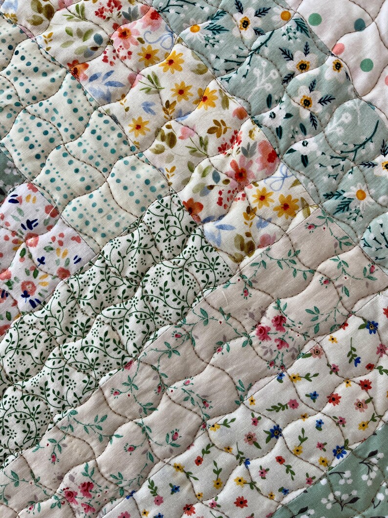 Pu&ograve; includere: Primo piano di un tessuto trapuntato con un design patchwork. La trapunta presenta vari motivi floreali nei toni del rosa, giallo, blu e verde su sfondo bianco. Le cuciture creano un motivo diagonale strutturato sul tessuto.