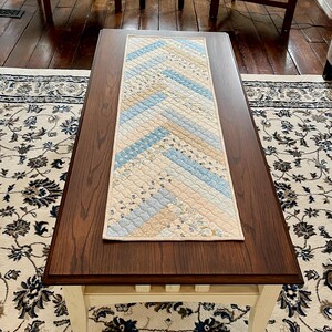 May include: A rectangular quilted table runner with a cream-coloured border. The centre features a patchwork design of light blue, cream, and floral patterned fabric in a chevron pattern. The runner is placed on a dark brown wooden coffee table.