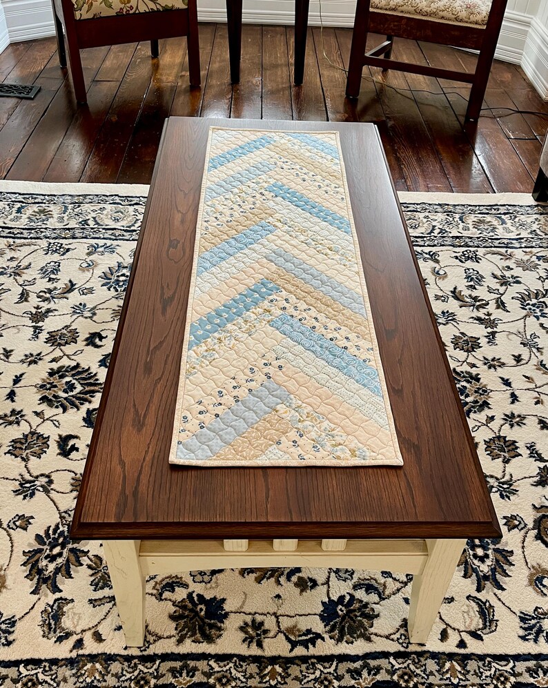 May include: A rectangular quilted table runner with a cream-coloured border. The centre features a patchwork design of light blue, cream, and floral patterned fabric in a chevron pattern. The runner is placed on a dark brown wooden coffee table.