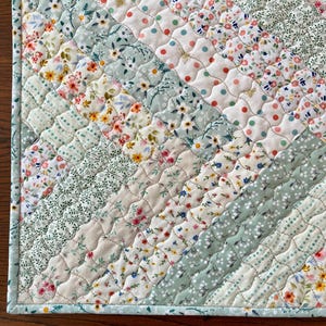 Pu&ograve; includere: Un sottopiatto trapuntato con un design patchwork. Il sottopiatto presenta file diagonali di tessuto in vari motivi e colori floreali, tra cui bianco, azzurro e beige. I bordi sono rifiniti con un bordo azzurro.