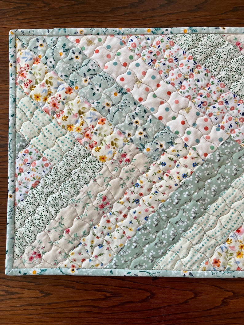 Pu&ograve; includere: Un sottopiatto trapuntato con un design patchwork. Il sottopiatto presenta file diagonali di tessuto in vari motivi e colori floreali, tra cui bianco, azzurro e beige. I bordi sono rifiniti con un bordo azzurro.