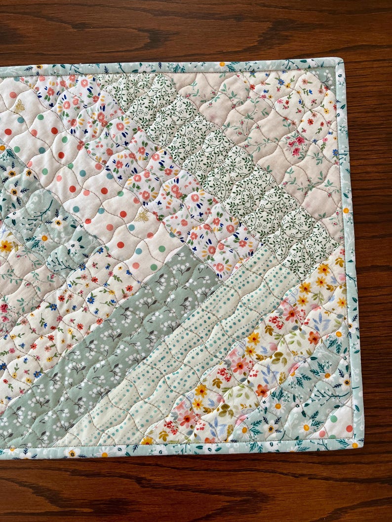 Pu&ograve; includere: Un sottopiatto trapuntato con un motivo diagonale di tessuti floreali e a pois in colori pastello. I tessuti includono bianco, azzurro e rosa con piccoli motivi floreali e a pois. I bordi sono rifiniti con un bordo azzurro.