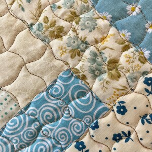 May include: Close-up of a quilted fabric with a patchwork design. The quilt features various floral patterns in shades of blue, cream, and green, with a light blue background. The stitching creates a diamond pattern.