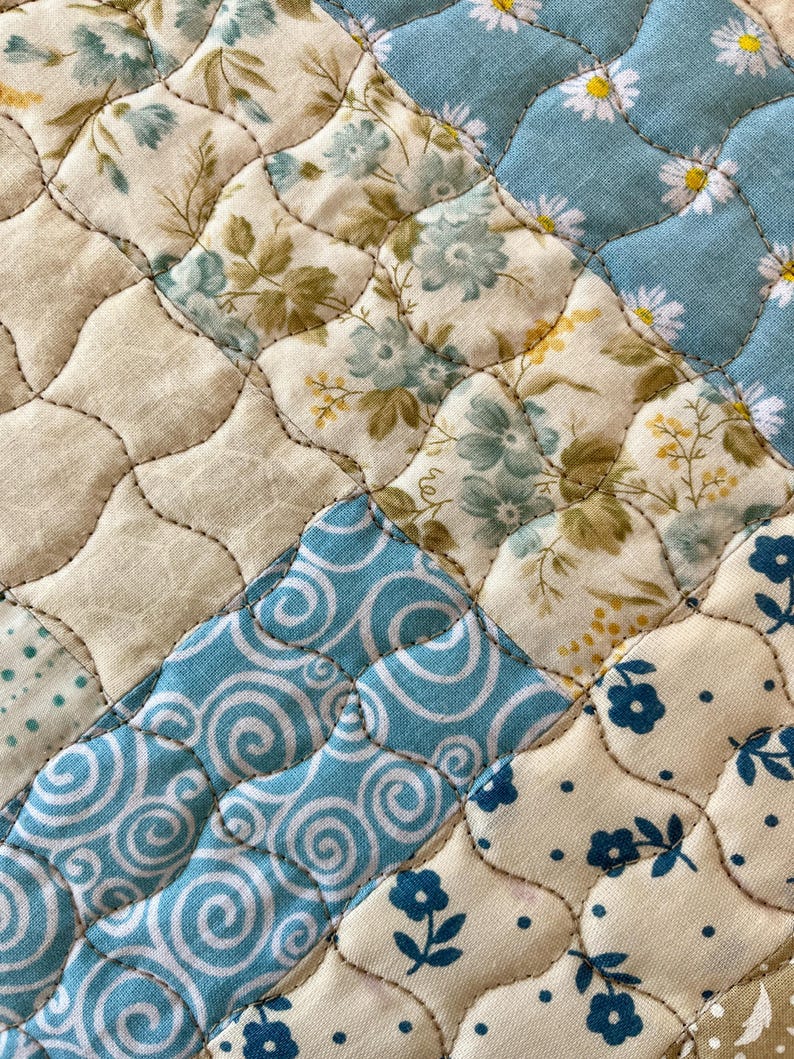May include: Close-up of a quilted fabric with a patchwork design. The quilt features various floral patterns in shades of blue, cream, and green, with a light blue background. The stitching creates a diamond pattern.