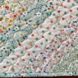 Pu&ograve; includere: Primo piano di un tessuto trapuntato con una variet&agrave; di motivi floreali in colori pastello. La trapunta presenta file diagonali di tessuti diversi, tra cui stampe floreali, pois e un motivo a forma di ape. I bordi sono rifiniti con un bordo azzurro.