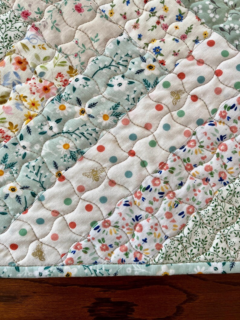 Pu&ograve; includere: Primo piano di un tessuto trapuntato con una variet&agrave; di motivi floreali in colori pastello. La trapunta presenta file diagonali di tessuti diversi, tra cui stampe floreali, pois e un motivo a forma di ape. I bordi sono rifiniti con un bordo azzurro.