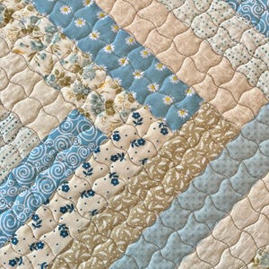 May include: A quilted placemat featuring a diagonal design of patterned fabrics. The fabrics include light blue with daisy prints, floral patterns, and solid beige and light blue. The placemat has a beige border and quilted stitching.