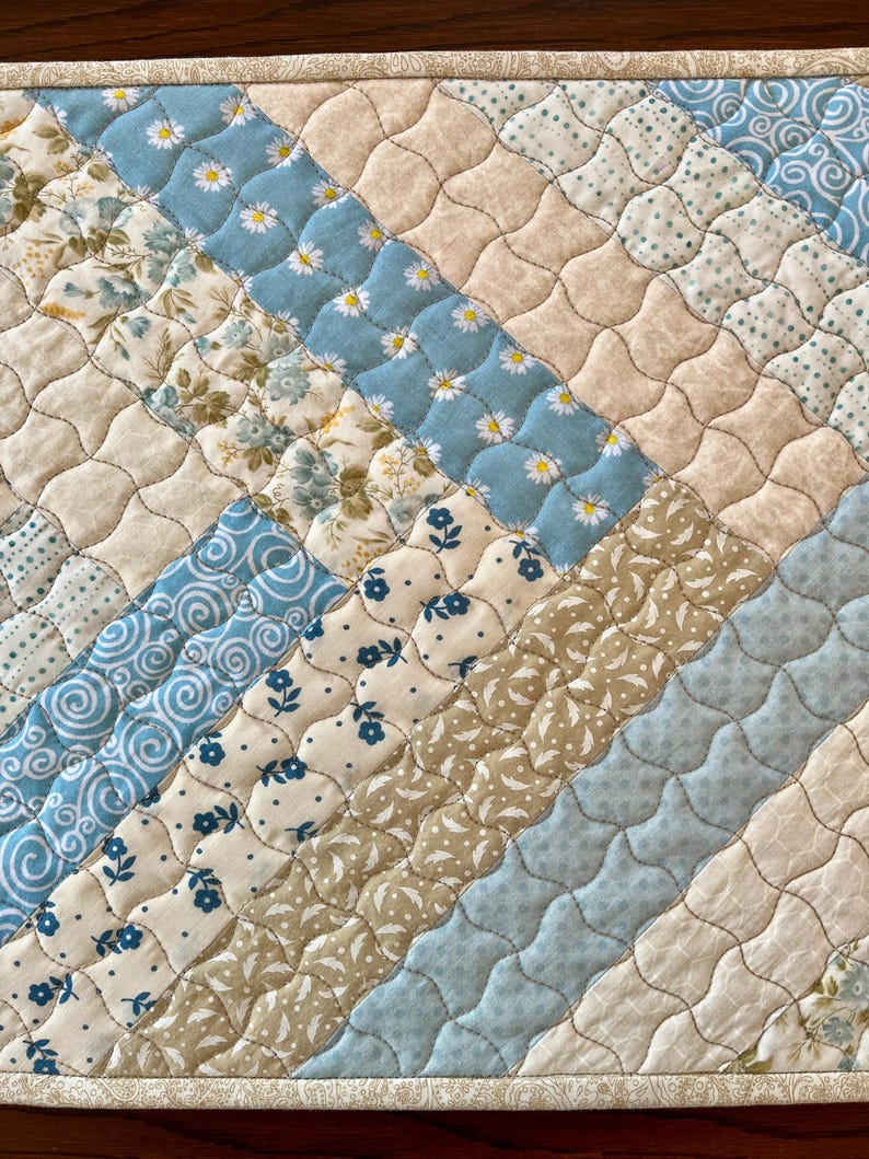May include: A quilted placemat featuring a diagonal design of patterned fabrics. The fabrics include light blue with daisy prints, floral patterns, and solid beige and light blue. The placemat has a beige border and quilted stitching.