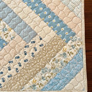 May include: A quilted placemat featuring a diagonal design of assorted fabrics. The pattern incorporates light blue, cream, and tan fabrics with floral and daisy motifs. The edges are finished with a patterned border.