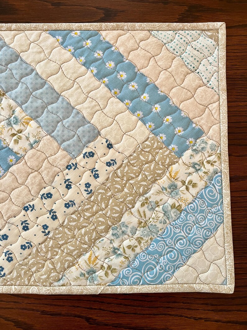 May include: A quilted placemat featuring a diagonal design of assorted fabrics. The pattern incorporates light blue, cream, and tan fabrics with floral and daisy motifs. The edges are finished with a patterned border.