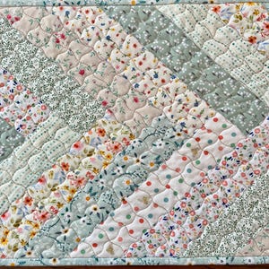 Pu&ograve; includere: Runner da tavolo trapuntato con un design patchwork. Il runner presenta strisce diagonali di vari motivi floreali e a pois in colori pastello, tra cui rosa, verde e bianco. I bordi sono rifiniti con un bordo azzurro.