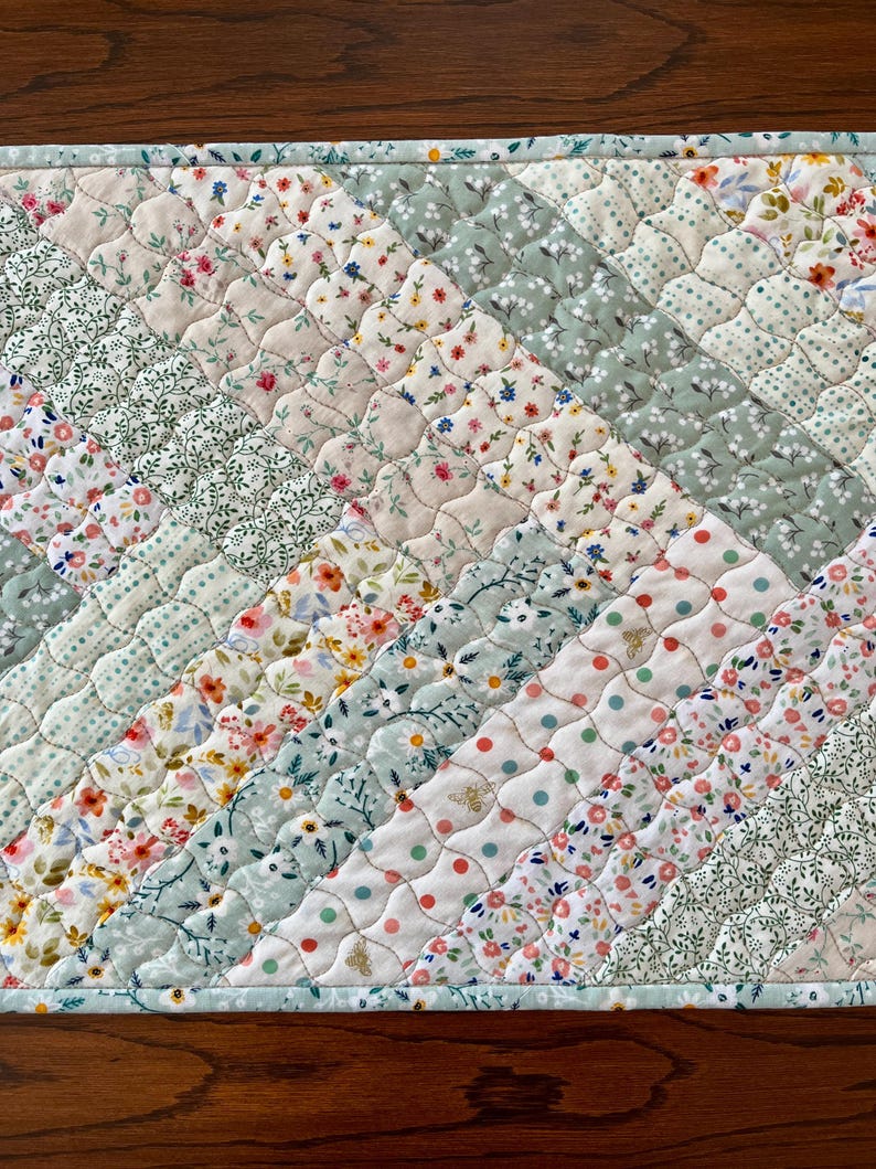 Pu&ograve; includere: Runner da tavolo trapuntato con un design patchwork. Il runner presenta strisce diagonali di vari motivi floreali e a pois in colori pastello, tra cui rosa, verde e bianco. I bordi sono rifiniti con un bordo azzurro.