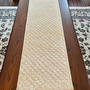 May include: A beige quilted table runner with a repeating pattern of small, connected shapes. The runner is rectangular and lies on a dark brown wooden table. The edges are neatly finished with a matching border. The runner is approximately 152 cm long.