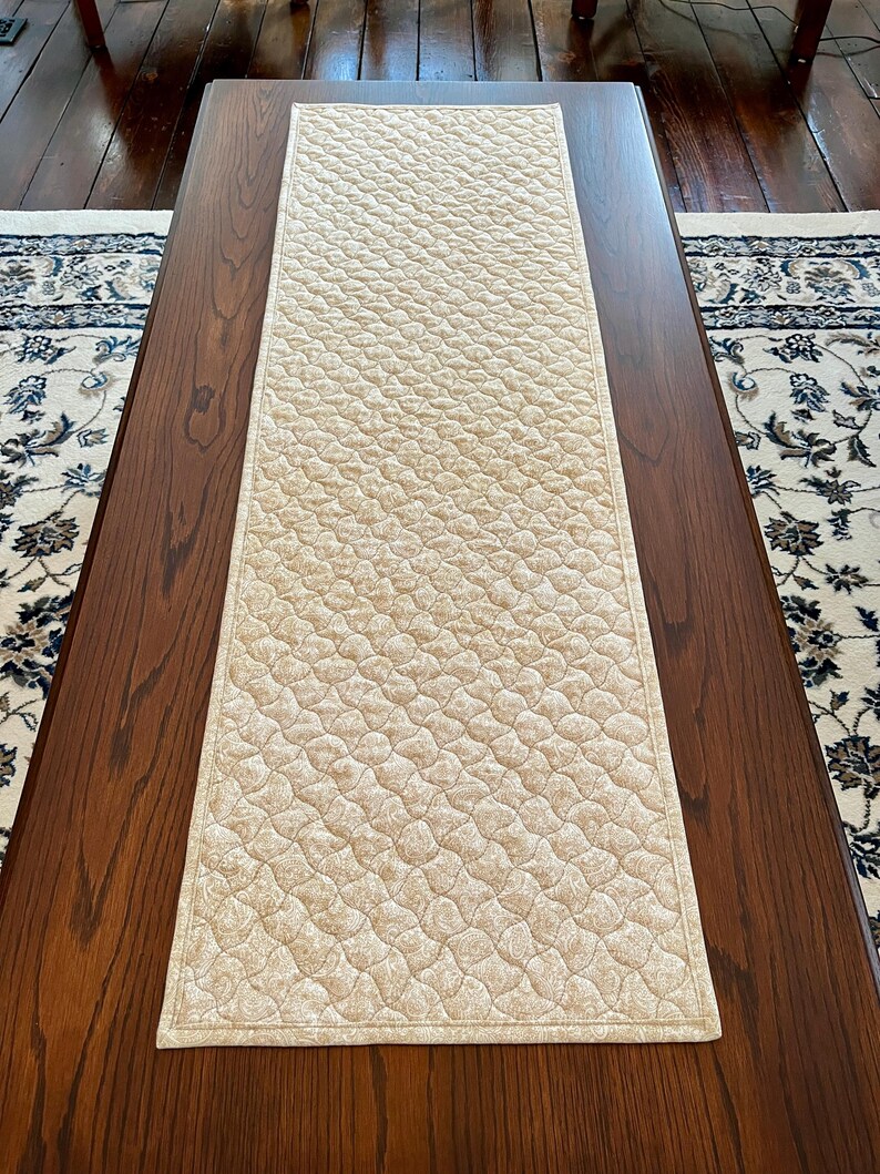 May include: A beige quilted table runner with a repeating pattern of small, connected shapes. The runner is rectangular and lies on a dark brown wooden table. The edges are neatly finished with a matching border. The runner is approximately 152 cm long.