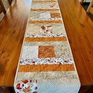 May include: A patchwork table runner in shades of orange, beige, and white, with floral and leaf patterns. The runner is placed on a wooden table. The runner's design is quilted.