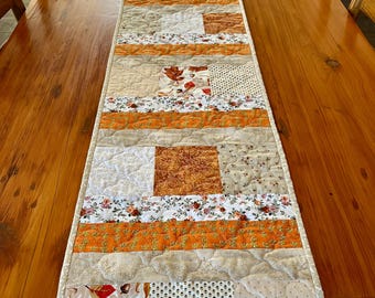 May include: A patchwork table runner in shades of orange, beige, and white, with floral and leaf patterns. The runner is placed on a wooden table. The runner's design is quilted.