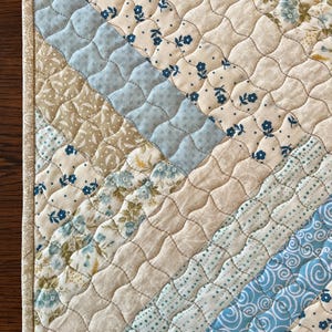 May include: A quilted placemat featuring a diagonal design in shades of light blue, beige, and floral patterns. The edges are trimmed with a patterned fabric, and the piece is detailed with intricate stitching. The design incorporates floral and geometric motifs.