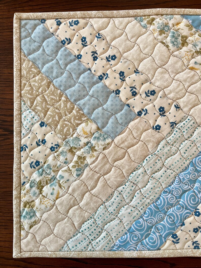 May include: A quilted placemat featuring a diagonal design in shades of light blue, beige, and floral patterns. The edges are trimmed with a patterned fabric, and the piece is detailed with intricate stitching. The design incorporates floral and geometric motifs.