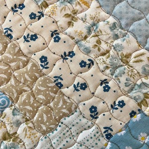 May include: Close-up of a quilted fabric with a patchwork design. The quilt features various floral patterns in shades of blue, beige, and brown. The stitching creates a textured, diamond-shaped pattern, adding depth and visual interest to the textile.