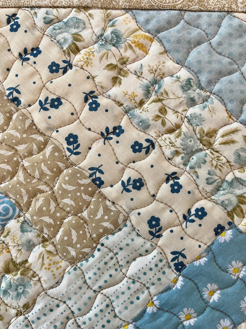 May include: Close-up of a quilted fabric with a patchwork design. The quilt features various floral patterns in shades of blue, beige, and brown. The stitching creates a textured, diamond-shaped pattern, adding depth and visual interest to the textile.