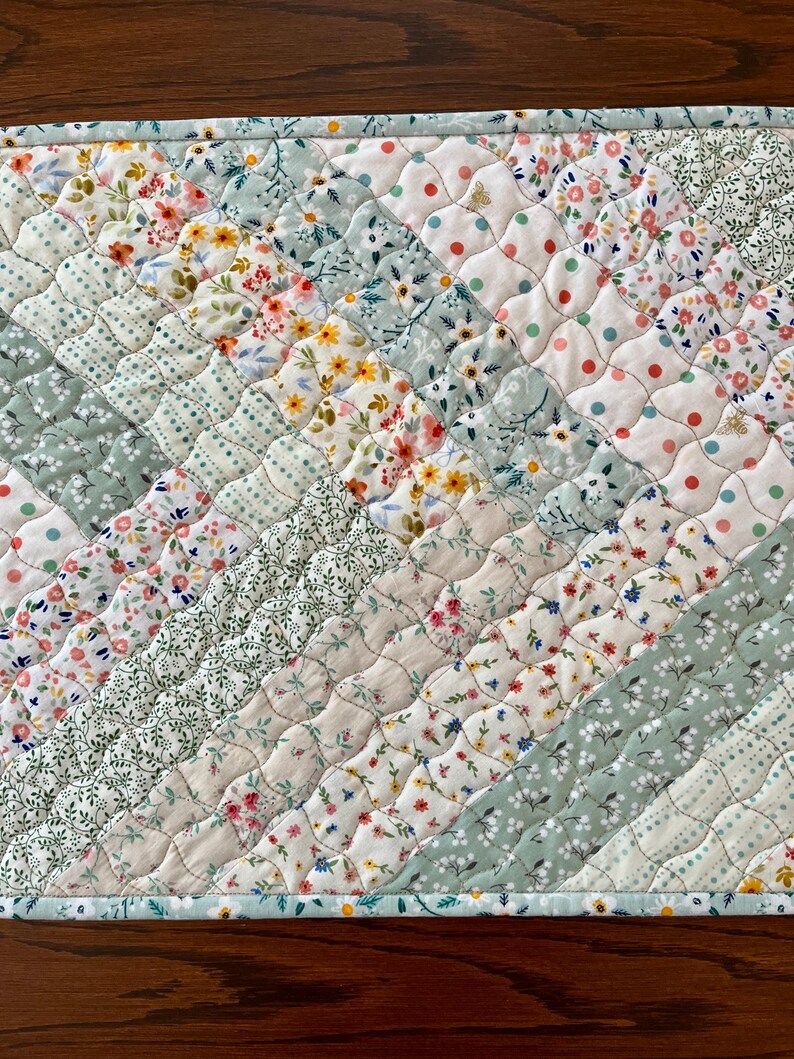 Pu&ograve; includere: Runner da tavolo trapuntato con un design patchwork. Il runner presenta vari motivi floreali e a pois in colori pastello, tra cui rosa, giallo e verde. Il motivo diagonale &egrave; cucito con un motivo a diamante. I bordi sono rifiniti con un bordo azzurro.