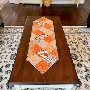 May include: A diamond-shaped quilted table runner featuring a patchwork design of orange, brown, and cream fabrics. The pattern includes various autumn-themed prints such as falling leaves, small acorns, and floral motifs arranged in a grid.