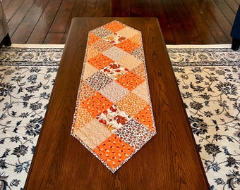 May include: A diamond-shaped quilted table runner featuring a patchwork design of orange, brown, and cream fabrics. The pattern includes various autumn-themed prints such as falling leaves, small acorns, and floral motifs arranged in a grid.