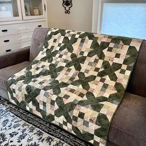 May include: A patchwork quilt with a star design in green, beige, and blue tones, placed on a brown sofa. The quilt's pattern is made up of intersecting shapes, giving it a textured look. This quilt is a decorative home accessory, perfect for adding warmth.