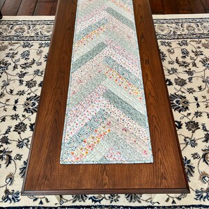 Pu&ograve; includere: Una tovaglia rettangolare trapuntata con un motivo a spina di pesce. La tovaglia presenta un design patchwork con varie strisce di tessuto floreale e tinta unita in colori pastello, tra cui rosa, blu e verde. La tovaglia &egrave; posizionata su un tavolino da caff&egrave; in legno marrone scuro.