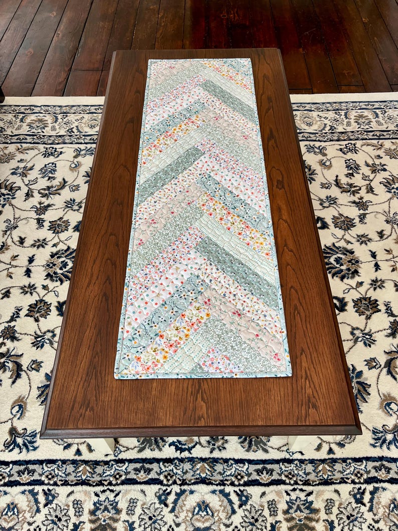 Pu&ograve; includere: Una tovaglia rettangolare trapuntata con un motivo a spina di pesce. La tovaglia presenta un design patchwork con varie strisce di tessuto floreale e tinta unita in colori pastello, tra cui rosa, blu e verde. La tovaglia &egrave; posizionata su un tavolino da caff&egrave; in legno marrone scuro.