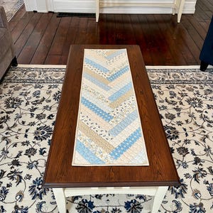 May include: A rectangular quilted table runner with a chevron pattern. The runner features alternating strips of light blue, beige, and floral patterned fabric. It is placed on a dark brown wooden coffee table, adding a decorative touch to the living room.