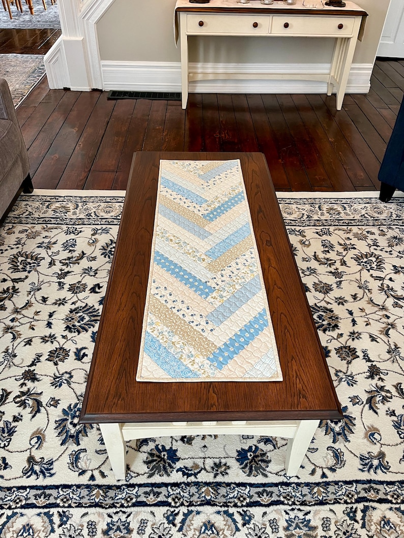May include: A rectangular quilted table runner with a chevron pattern. The runner features alternating strips of light blue, beige, and floral patterned fabric. It is placed on a dark brown wooden coffee table, adding a decorative touch to the living room.