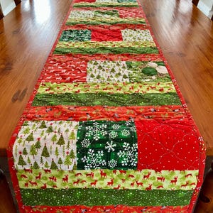 May include: A festive Christmas table runner with a patchwork design. The runner features red, green, and white fabrics with patterns of reindeer, snowflakes, and Christmas trees. The runner is displayed on a wooden table.