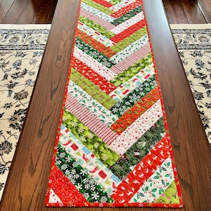 May include: A festive Christmas table runner with a chevron design. The runner is made from red, green, and white patterned fabrics, featuring festive motifs like snowflakes and candy canes. It is rectangular and rests on a wooden coffee table.