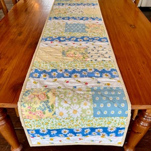 Peut inclure: Un chemin de table en patchwork avec un motif floral dans les tons de bleu, jaune et blanc. Le chemin de table est matelassé et a une bordure blanche.