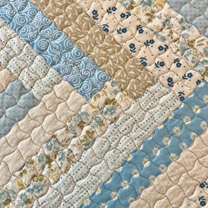 May include: A quilted placemat with a diagonal pattern of various fabrics. The fabrics include light blue with white daisy patterns, cream with small blue floral prints, and tan with floral patterns. The edges are finished with a patterned border.